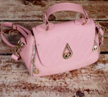 A pink handbag with a quilted pattern, gold hardware, and a distinctive raindrop-shaped embellishment on the front. It rests on a textured wooden surface, giving a rustic contrast to the bag's elegant design.