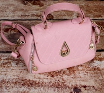 A pink handbag with a quilted pattern, gold hardware, and a distinctive raindrop-shaped embellishment on the front. It rests on a textured wooden surface, giving a rustic contrast to the bag's elegant design.