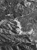 An aerial view of a mountainous landscape, displaying a natural array of jagged peaks and valleys. The terrain is rugged with a combination of steep slopes and ridges interspersed with flatter areas, possibly representing terraces or fields. Shadows and light create a high contrast effect, highlighting the topographical texture.