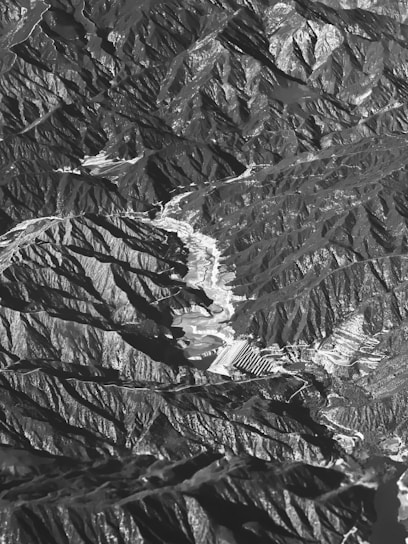 An aerial view of a mountainous landscape, displaying a natural array of jagged peaks and valleys. The terrain is rugged with a combination of steep slopes and ridges interspersed with flatter areas, possibly representing terraces or fields. Shadows and light create a high contrast effect, highlighting the topographical texture.