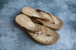 A pair of beige flip-flops with decorative floral patterns on the footbed. They are placed on a textured grey surface. The sandals have thick soles, and there are metal embellishments on the straps.