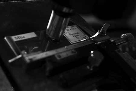 A black and white close-up of a microscope setup with a slide labeled 'Histology' and 'Mammal Heart'. The slide is placed under the microscope lens, and the image prominently features the mechanical parts of the microscope.