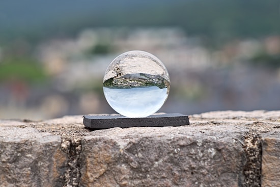 A clear crystal ball is positioned on a small rectangular black stand, placed on a rough stone surface. The background shows an out-of-focus landscape with greenery, mountains, and buildings, which are inverted and clearly visible through the crystal ball.
