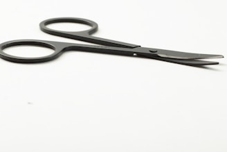 A pair of small, black scissors with two finger holes, placed on a smooth, white surface. The blades are slightly open and angled away from the viewer.