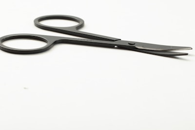 A pair of small, black scissors with two finger holes, placed on a smooth, white surface. The blades are slightly open and angled away from the viewer.