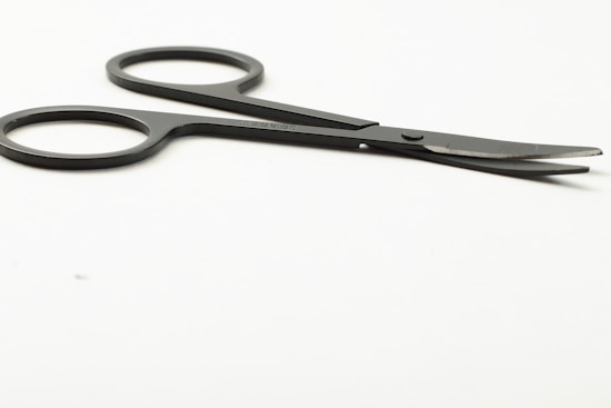 A pair of small, black scissors with two finger holes, placed on a smooth, white surface. The blades are slightly open and angled away from the viewer.