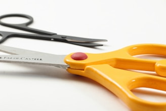 A pair of large, orange-handled scissors with a red button is in the foreground, with smaller black scissors partially visible in the background. The brand name on the blade reads 'Faber-Castell'. The scene is set against a plain white background, highlighting the bright color of the handles.