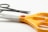 A pair of large, orange-handled scissors with a red button is in the foreground, with smaller black scissors partially visible in the background. The brand name on the blade reads 'Faber-Castell'. The scene is set against a plain white background, highlighting the bright color of the handles.