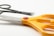 A pair of large, orange-handled scissors with a red button is in the foreground, with smaller black scissors partially visible in the background. The brand name on the blade reads 'Faber-Castell'. The scene is set against a plain white background, highlighting the bright color of the handles.