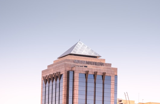 A modern office building with a geometric, pyramid-shaped top. The architecture features reflective glass windows and a beige stone exterior. The name 'Mutua Madrileña' is displayed near the top.