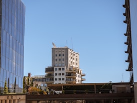 The image features a cityscape with a tall beige building positioned centrally, surrounded by modern glass structures. The sky is clear blue, and the urban scene includes balconies, greenery, and antennas atop the buildings.