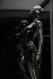 A sleek and reflective humanoid robot stands against a dark background. The robot's surface is glossy and metallic, giving it a futuristic and high-tech appearance. The structure of the robot is humanoid with defined limbs and a head, and it appears to be contemplating or observing something. The dark and reflective environment around the robot emphasizes its metallic features.
