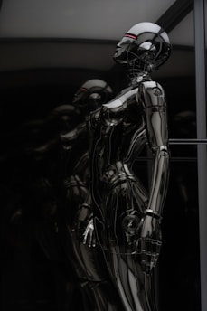 A sleek and reflective humanoid robot stands against a dark background. The robot's surface is glossy and metallic, giving it a futuristic and high-tech appearance. The structure of the robot is humanoid with defined limbs and a head, and it appears to be contemplating or observing something. The dark and reflective environment around the robot emphasizes its metallic features.