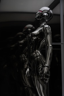 A sleek and reflective humanoid robot stands against a dark background. The robot's surface is glossy and metallic, giving it a futuristic and high-tech appearance. The structure of the robot is humanoid with defined limbs and a head, and it appears to be contemplating or observing something. The dark and reflective environment around the robot emphasizes its metallic features.