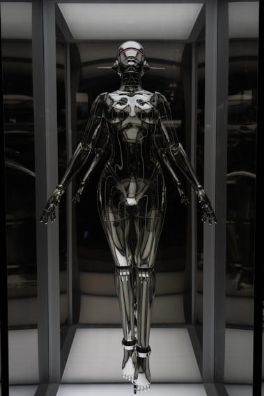 A humanoid robot with a futuristic design stands in an illuminated display case. The robot has a sleek, metallic body with articulated joints and a smooth, reflective surface. Its head is shaped with minimalistic features, including a visor-like element covering where eyes would be. The robot is positioned upright with arms slightly extended to the sides.