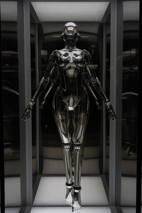A humanoid robot with a futuristic design stands in an illuminated display case. The robot has a sleek, metallic body with articulated joints and a smooth, reflective surface. Its head is shaped with minimalistic features, including a visor-like element covering where eyes would be. The robot is positioned upright with arms slightly extended to the sides.