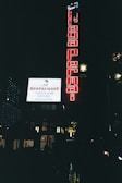 A nighttime urban scene featuring illuminated signs on a building. A large red vertical sign displays the words 'Braga Permai,' and a bright white horizontal sign advertises a restaurant with the words 'Chocolatier,' 'Pâtisseries,' and 'Boulangers.' The scene is dark, with some ambient light and reflections on wet streets below.