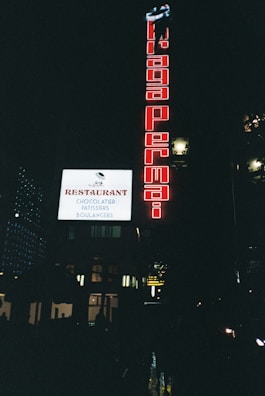 A nighttime urban scene featuring illuminated signs on a building. A large red vertical sign displays the words 'Braga Permai,' and a bright white horizontal sign advertises a restaurant with the words 'Chocolatier,' 'Pâtisseries,' and 'Boulangers.' The scene is dark, with some ambient light and reflections on wet streets below.