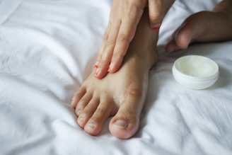 Close-up of a person applying creamy body butter to their feet in a cozy bathroom setting.
