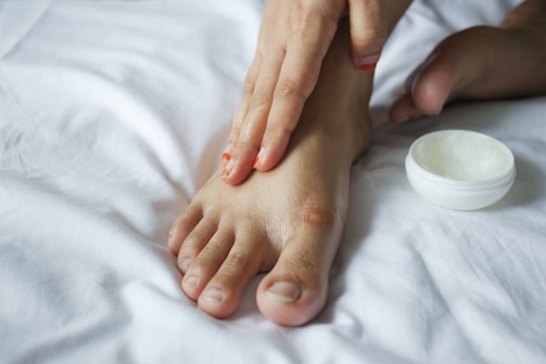 A person's hand is applying cream to their foot on a white bed. The scene captures a moment of personal care and relaxation.
