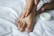 A person's hand is applying cream to their foot on a white bed. The scene captures a moment of personal care and relaxation.