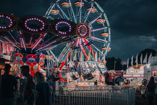 A lively fairground scene at night featuring a brightly lit Ferris wheel in the background and a colorful swing ride in the foreground. A few people are visible near the rides, and the atmosphere is festive with illuminated carnival booths lining the scene.