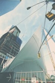 A modern architectural structure with sleek, white, ribbed design elements, possibly part of a larger building project. Surrounding it are high-rise buildings and a construction crane. The scene includes a traffic signal and a construction site sign indicating Gate 3H. The sky is mostly clear with light clouds.