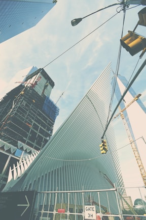 A modern architectural structure with sleek, white, ribbed design elements, possibly part of a larger building project. Surrounding it are high-rise buildings and a construction crane. The scene includes a traffic signal and a construction site sign indicating Gate 3H. The sky is mostly clear with light clouds.
