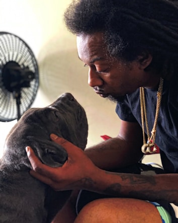 A person with dreadlocks gently holds and looks into the eyes of a large gray dog. The scene conveys a sense of affection and a close bond between the two. A fan is visible in the background, suggesting a casual indoor setting.