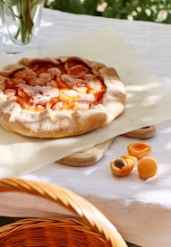 A freshly baked fruit tart with a golden crust, topped with halved apricots, and lightly dusted with powdered sugar. It rests on parchment paper on a wooden cutting board placed on a white tablecloth. Nearby, there are a few whole and halved apricots. A wicker basket is partially visible in the foreground, and a vase with green stems is slightly visible in the background.