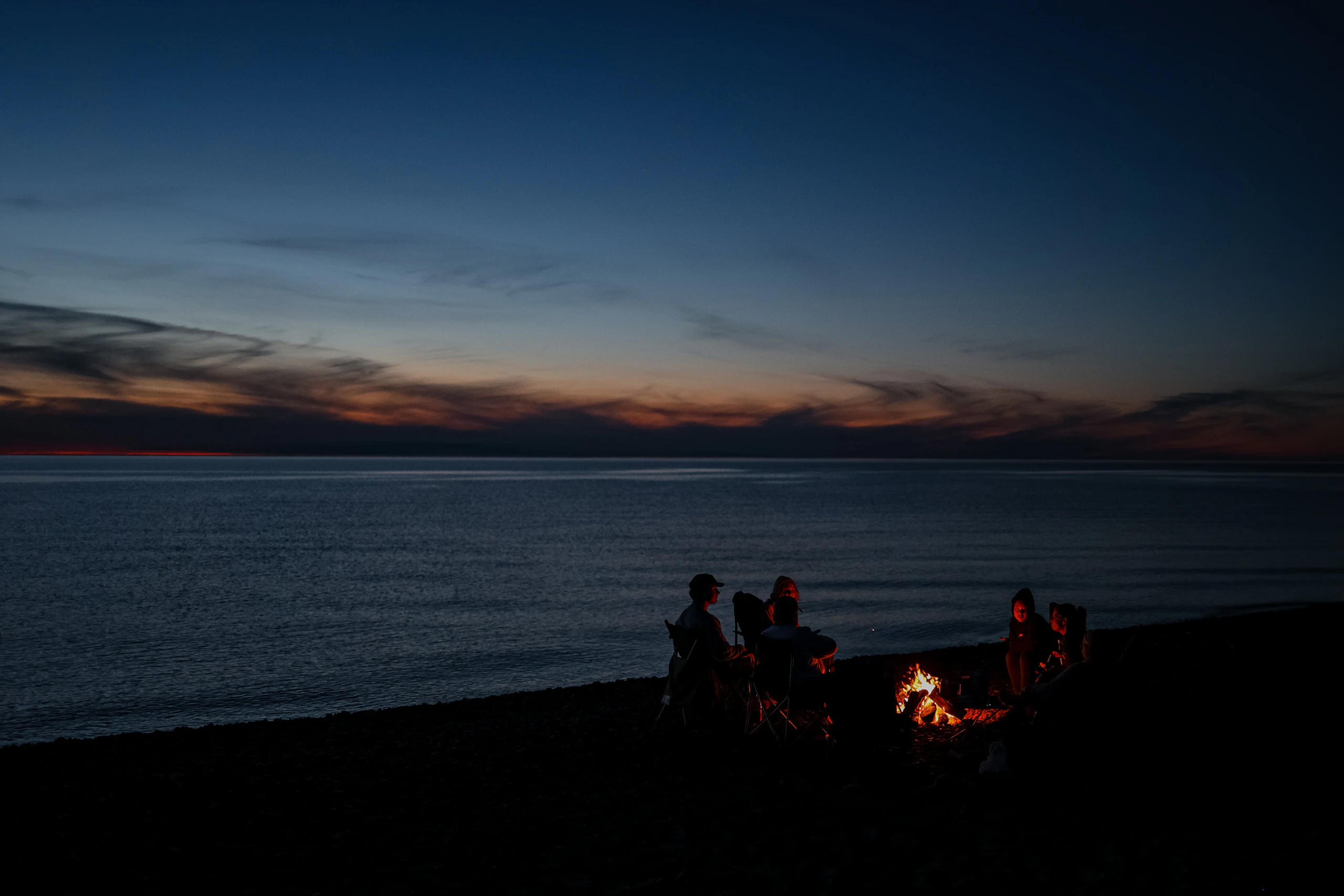 A small group around a beach fire at dusk beside calm water