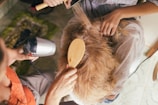 A person is having their hair styled. Another person uses a round brush and a hairdryer on the blonde hair while wearing an orange top. The person receiving the styling is sitting on a chair, covered with a towel.