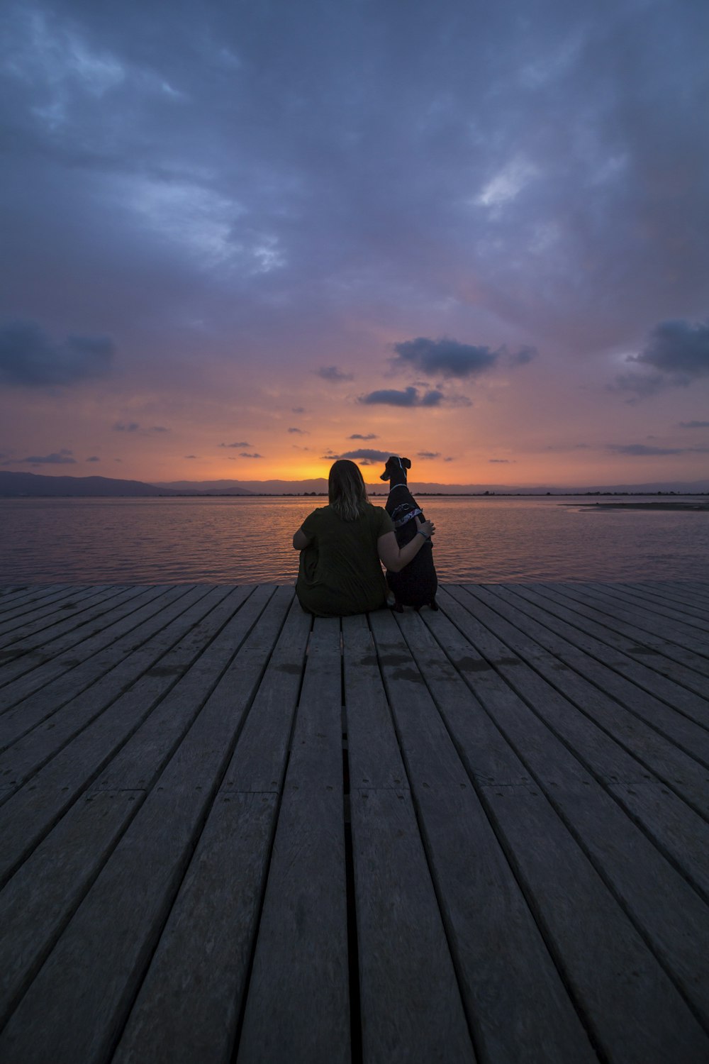 woman in black jacket sitting on wooden dock during sunset