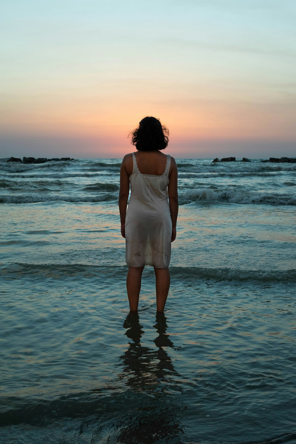 Frau in weißem Kleid steht bei Sonnenuntergang am Meeresufer