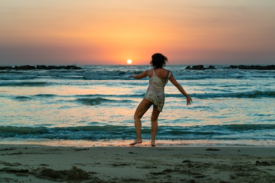 A person is dancing or playfully moving on the beach, facing the setting sun. The sky is painted in warm hues of orange and pink, with gentle waves lapping the shore.