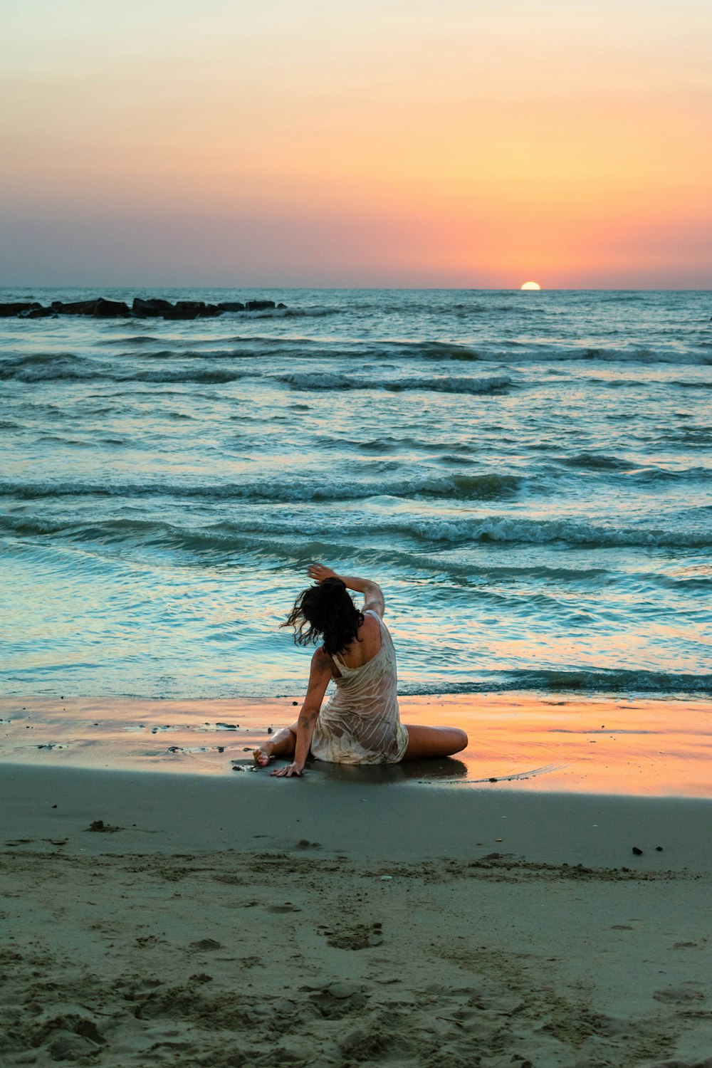 Mujer en camiseta blanca sin mangas sentada en la playa durante la puesta del sol