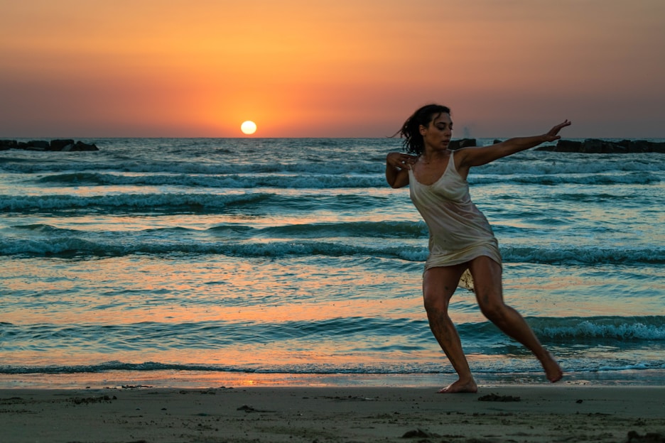 Full-screen video still showing women moving gracefully on the beach with sunlight highlighting fitness outfits in black, white, gold, ocean blue, and nude tones