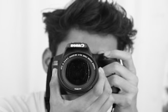 A person holds a Canon camera close to their face, focusing on the lens as if taking a photograph. The image is in black and white, adding a classic, timeless feel.