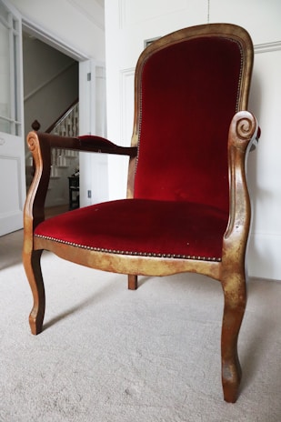 A vintage wooden chair with intricate carved details on the arms and a high backrest. The chair is upholstered in rich red velvet, and it sits on a light-colored carpet in a well-lit room.