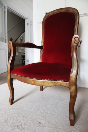 A vintage wooden chair with intricate carved details on the arms and a high backrest. The chair is upholstered in rich red velvet, and it sits on a light-colored carpet in a well-lit room.