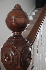 A close-up view of a decorative wooden staircase newel post featuring detailed carvings and a polished finish. The wooden post contrasts with the painted white staircase railing and steps in the background, providing an elegant, classic look.