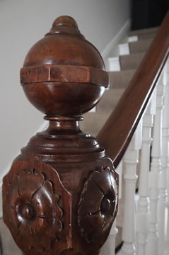 A close-up view of a decorative wooden staircase newel post featuring detailed carvings and a polished finish. The wooden post contrasts with the painted white staircase railing and steps in the background, providing an elegant, classic look.