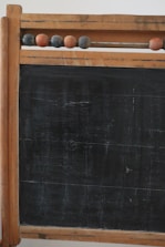 A vintage wooden-framed chalkboard with an abacus at the top. The abacus consists of several wooden beads strung on a metal rod. The chalkboard surface shows signs of wear and age, with visible scratches and marks, giving it an antique look.