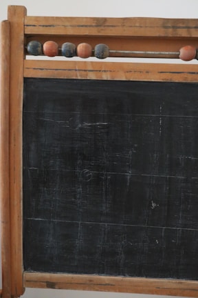 A vintage wooden-framed chalkboard with an abacus at the top. The abacus consists of several wooden beads strung on a metal rod. The chalkboard surface shows signs of wear and age, with visible scratches and marks, giving it an antique look.