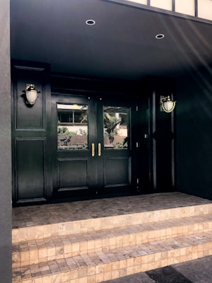 A dark, elegant double door entrance with glass panels is flanked by two wall-mounted lamps. The entrance is approached via a short flight of light-colored stone steps. Reflections of outdoor vegetation are visible in the glass panels, and the overall structure is framed by a dark facade.