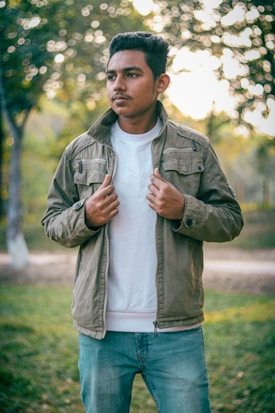 A man is standing outdoors wearing a khaki jacket over a white shirt and blue jeans. The background consists of blurred greenery and trees with dappled sunlight filtering through the leaves, creating a warm and peaceful atmosphere. The man has a relaxed yet thoughtful expression.