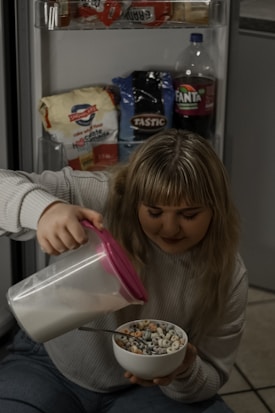 A woman is sitting on the floor in front of an open refrigerator, pouring milk from a container with a pink lid into a bowl of colorful cereal. The refrigerator door holds various food items including bread, rice, and a bottle of Fanta. She has long, light-colored hair and is wearing a light-colored sweater and jeans.