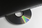 A CD partially inserted into a sleek, modern laptop. The disc displays a spectrum of colors due to light reflection, and the laptop is minimalist in design with visible ports on the side.