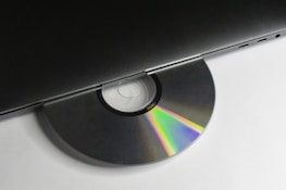 A CD partially inserted into a sleek, modern laptop. The disc displays a spectrum of colors due to light reflection, and the laptop is minimalist in design with visible ports on the side.