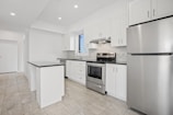 A modern kitchen featuring white cabinetry, stainless steel appliances, and a sleek black countertop. The room is well-lit with recessed lighting and has large floor tiles that add to the spacious feel. A small island provides additional workspace.